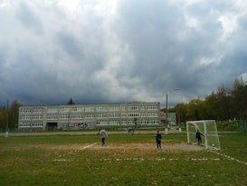 A school building under a cloudy sky with a football field in the foreground. Several people are present on the field, including children playing near a goalpost. The setting appears to be outdoors within a schoolyard environment surrounded by trees.