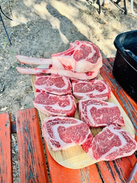 Several cuts of raw red meat are arranged on a wooden board placed on top of a rustic red-painted picnic table. The setting appears to be outdoors, with dirt and some leaves visible around the table.