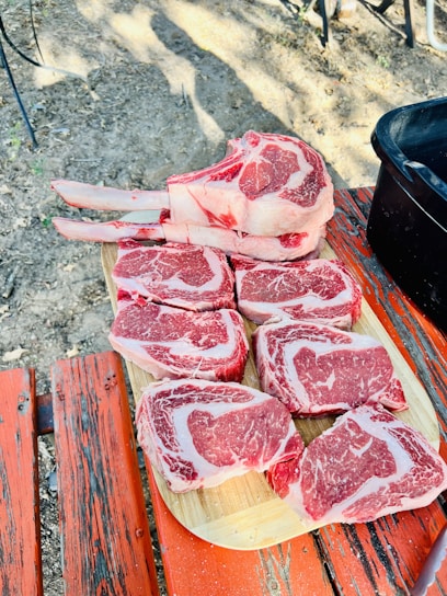 Several cuts of raw red meat are arranged on a wooden board placed on top of a rustic red-painted picnic table. The setting appears to be outdoors, with dirt and some leaves visible around the table.