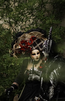 Two individuals are adorned in elaborate, theatrical costumes. They wear ornate, decorated headpieces, and their faces are painted with dramatic makeup. The attire includes intricate details and patterns with a dark, antique style, set against a backdrop of lush greenery.