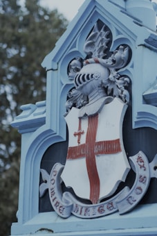 A detailed carving features a medieval knight&rsquo;s helmet atop a shield, which is emblazoned with a cross and a sword. The decoration includes ornate elements such as leaves and intricate patterns. The structure is predominantly a pale blue color, with the shield having a red cross. The inscription on a ribbon beneath the shield reads 'DOMINE DIRIGE NOS' in a Gothic font. In the background, there are trees visible, indicating an outdoor setting.