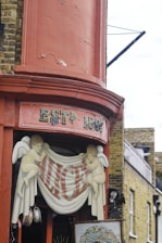 A decorative corner with aged red paint and the sign 'ESTD 1862'. Two painted cherub figures hold a banner with the name 'ALICE'S'. The background features traditional brickwork and architectural details typical of an older building.