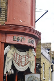 A decorative corner with aged red paint and the sign 'ESTD 1862'. Two painted cherub figures hold a banner with the name 'ALICE'S'. The background features traditional brickwork and architectural details typical of an older building.