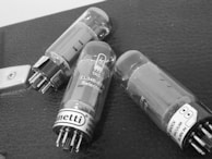 Three vacuum tubes are placed on a textured surface, possibly a piece of equipment or an amplifier. Each tube has metallic pins protruding at one end and clear glass encasing with visible internal structures. Labels with brand and specifications are attached near the pin area.