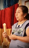 A woman is holding a dried corn cob, wearing a patterned apron with traditional embroidery. Behind her, skeins of colorful yarns in shades of blue, red, and yellow hang on a wall. The setting appears to be artisanal, possibly related to weaving or textile work.