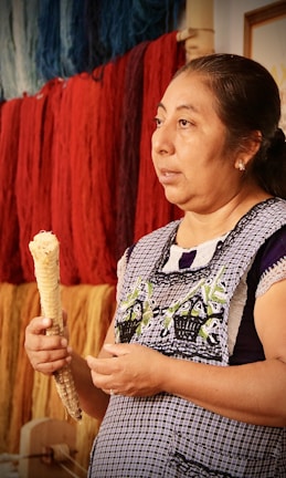 A woman is holding a dried corn cob, wearing a patterned apron with traditional embroidery. Behind her, skeins of colorful yarns in shades of blue, red, and yellow hang on a wall. The setting appears to be artisanal, possibly related to weaving or textile work.