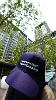 A person wearing a navy blue cap with the phrase 'Keep your head cool while trading' is positioned in the foreground. The background features a high-rise residential building surrounded by green trees and a bright sky.