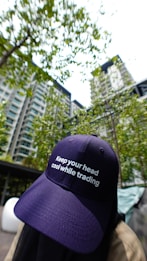 A person wearing a navy blue cap with the phrase 'Keep your head cool while trading' is positioned in the foreground. The background features a high-rise residential building surrounded by green trees and a bright sky.