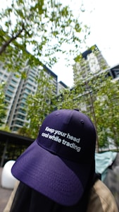 A person wearing a navy blue cap with the phrase 'Keep your head cool while trading' is positioned in the foreground. The background features a high-rise residential building surrounded by green trees and a bright sky.