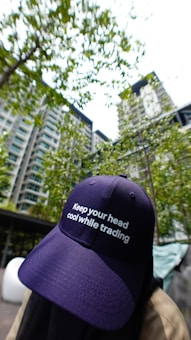 A person wearing a navy blue cap with the phrase 'Keep your head cool while trading' is positioned in the foreground. The background features a high-rise residential building surrounded by green trees and a bright sky.