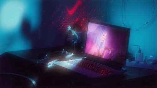 A dimly lit room with an ambient blue glow features a laptop displaying an image of a woman with neon pink hair. The scene includes a game controller and some scattered desk items, while red laser lights decorate the background, adding a futuristic feel.