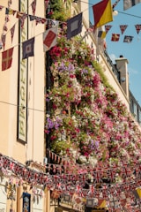 A vertical garden on a building facade is covered in a vibrant and dense display of flowers in purple, pink, red, white, and green shades. Multiple strings of UK-themed flags and other colorful pennants are strung across the street in festive decoration. The building features a light beige exterior with several decorative elements, including signage.