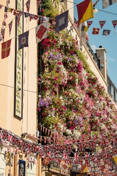 A vertical garden on a building facade is covered in a vibrant and dense display of flowers in purple, pink, red, white, and green shades. Multiple strings of UK-themed flags and other colorful pennants are strung across the street in festive decoration. The building features a light beige exterior with several decorative elements, including signage.
