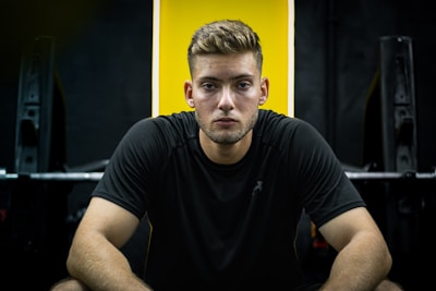 A man with a focused expression is sitting in a gym setting, wearing a black T-shirt. The background includes gym equipment and a bright yellow wall panel.