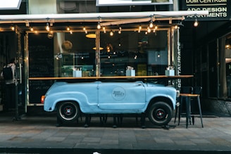 A creative outdoor cafe setup features a customized bar or counter crafted from a repurposed vintage car body, painted in light blue. The front of the car forms the base, while a wooden countertop rests on top. Several bar stools are positioned around the counter. Strings of warm lights hang above, adding ambiance. The cafe's modern and eclectic facade is visible in the background.