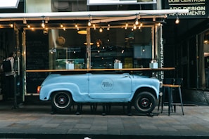 A creative outdoor cafe setup features a customized bar or counter crafted from a repurposed vintage car body, painted in light blue. The front of the car forms the base, while a wooden countertop rests on top. Several bar stools are positioned around the counter. Strings of warm lights hang above, adding ambiance. The cafe's modern and eclectic facade is visible in the background.