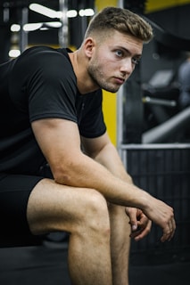 A young man with a focused expression is sitting with his elbows resting on his knees in a gym setting. He is wearing a black t-shirt and shorts, with exercise equipment visible in the background.