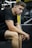A young man with a focused expression is sitting with his elbows resting on his knees in a gym setting. He is wearing a black t-shirt and shorts, with exercise equipment visible in the background.