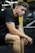 A young man with a focused expression is sitting with his elbows resting on his knees in a gym setting. He is wearing a black t-shirt and shorts, with exercise equipment visible in the background.
