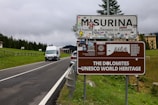 A road sign for Misurina and the Dolomites UNESCO World Heritage site is positioned next to a rural road surrounded by greenery. Various stickers and logos are attached to the sign, and a white van is driving toward it. Behind the sign, a line of trees and a large building can be seen under a cloudy sky.