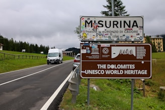 A road sign for Misurina and the Dolomites UNESCO World Heritage site is positioned next to a rural road surrounded by greenery. Various stickers and logos are attached to the sign, and a white van is driving toward it. Behind the sign, a line of trees and a large building can be seen under a cloudy sky.