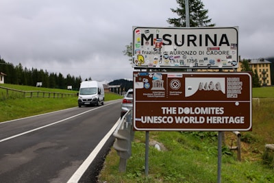 A road sign for Misurina and the Dolomites UNESCO World Heritage site is positioned next to a rural road surrounded by greenery. Various stickers and logos are attached to the sign, and a white van is driving toward it. Behind the sign, a line of trees and a large building can be seen under a cloudy sky.
