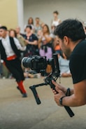 A photographer is holding a professional camera mounted on a stabilizer, capturing a scene with a blurred crowd in the background. A person in motion, dressed in traditional clothing, appears in the background, adding a dynamic element to the composition.