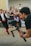 A photographer is holding a professional camera mounted on a stabilizer, capturing a scene with a blurred crowd in the background. A person in motion, dressed in traditional clothing, appears in the background, adding a dynamic element to the composition.