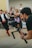 A photographer is holding a professional camera mounted on a stabilizer, capturing a scene with a blurred crowd in the background. A person in motion, dressed in traditional clothing, appears in the background, adding a dynamic element to the composition.
