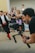 A photographer is holding a professional camera mounted on a stabilizer, capturing a scene with a blurred crowd in the background. A person in motion, dressed in traditional clothing, appears in the background, adding a dynamic element to the composition.