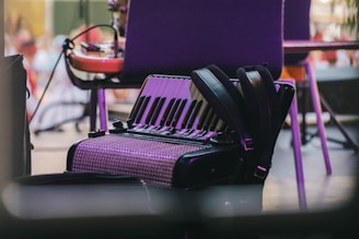 An accordion rests on the floor beside a chair, surrounded by musical equipment. The instrument's shiny keys and patterned body are highlighted under soft lighting, suggesting a relaxed setting.
