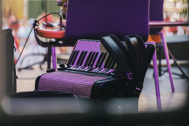 An accordion rests on the floor beside a chair, surrounded by musical equipment. The instrument's shiny keys and patterned body are highlighted under soft lighting, suggesting a relaxed setting.
