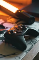 A video game controller rests on a patterned surface beside a glowing orange light source, creating a warm ambiance. The controller has a sleek, black design with joysticks and buttons, reflecting some of the orange light.