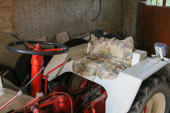 A vintage tractor is parked indoors, featuring a steering wheel and a seat with a floral cushion. The tractor is predominantly white with red accents, visible in the mechanical components and steering column. It is situated in a dimly lit space with concrete walls and a wooden door.