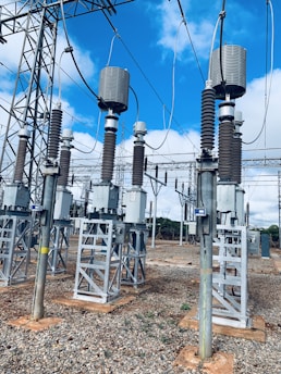 A high-voltage electrical substation with metal structures and transformers, set against a backdrop of a clear blue sky with some clouds. The substation components are mounted on concrete bases surrounded by gravel. Several large insulators and metal towers support the electrical equipment and power cables.