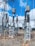 A high-voltage electrical substation with metal structures and transformers, set against a backdrop of a clear blue sky with some clouds. The substation components are mounted on concrete bases surrounded by gravel. Several large insulators and metal towers support the electrical equipment and power cables.