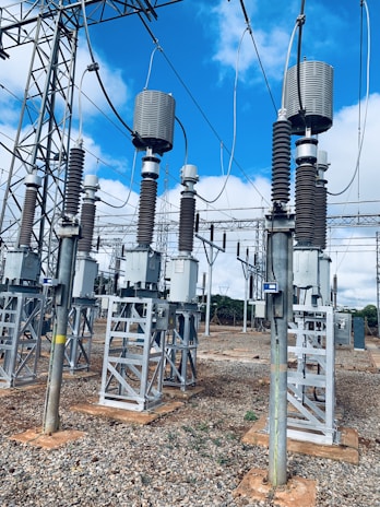 A high-voltage electrical substation with metal structures and transformers, set against a backdrop of a clear blue sky with some clouds. The substation components are mounted on concrete bases surrounded by gravel. Several large insulators and metal towers support the electrical equipment and power cables.