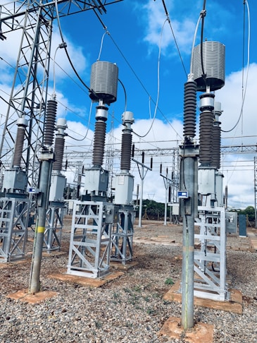 A high-voltage electrical substation with metal structures and transformers, set against a backdrop of a clear blue sky with some clouds. The substation components are mounted on concrete bases surrounded by gravel. Several large insulators and metal towers support the electrical equipment and power cables.