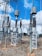 A high-voltage electrical substation with metal structures and transformers, set against a backdrop of a clear blue sky with some clouds. The substation components are mounted on concrete bases surrounded by gravel. Several large insulators and metal towers support the electrical equipment and power cables.
