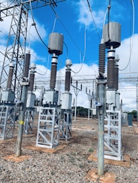 A high-voltage electrical substation with metal structures and transformers, set against a backdrop of a clear blue sky with some clouds. The substation components are mounted on concrete bases surrounded by gravel. Several large insulators and metal towers support the electrical equipment and power cables.