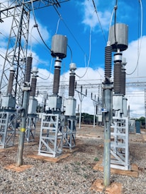 A high-voltage electrical substation with metal structures and transformers, set against a backdrop of a clear blue sky with some clouds. The substation components are mounted on concrete bases surrounded by gravel. Several large insulators and metal towers support the electrical equipment and power cables.