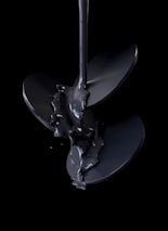 A stream of glossy black liquid pours over a shiny, metallic propeller, set against a dark background. The liquid appears thick and viscous, creating a reflective surface that highlights the curves and edges of the blades.