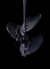 A stream of glossy black liquid pours over a shiny, metallic propeller, set against a dark background. The liquid appears thick and viscous, creating a reflective surface that highlights the curves and edges of the blades.