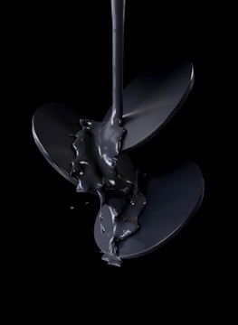A stream of glossy black liquid pours over a shiny, metallic propeller, set against a dark background. The liquid appears thick and viscous, creating a reflective surface that highlights the curves and edges of the blades.