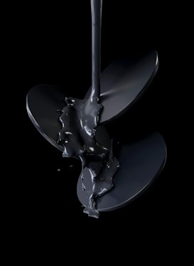 A stream of glossy black liquid pours over a shiny, metallic propeller, set against a dark background. The liquid appears thick and viscous, creating a reflective surface that highlights the curves and edges of the blades.