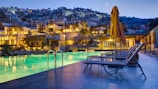 A luxurious outdoor area featuring a large swimming pool surrounded by lounge chairs and umbrellas. In the background, there are multiple levels of warmly lit, modern hotel buildings nestled into a hillside. The sky is a deepening blue with a crescent moon visible, adding to the serene atmosphere.