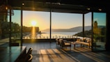 A peaceful room with a large glass wall that opens up to a breathtaking view of a sunset over a tranquil lake. The warm glow of the setting sun casts long shadows across the floor. Comfortable outdoor seating with cushions is positioned for enjoying the scenery. The interior includes a wooden dining table and chairs, with a cozy and serene atmosphere.