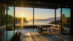 A peaceful room with a large glass wall that opens up to a breathtaking view of a sunset over a tranquil lake. The warm glow of the setting sun casts long shadows across the floor. Comfortable outdoor seating with cushions is positioned for enjoying the scenery. The interior includes a wooden dining table and chairs, with a cozy and serene atmosphere.