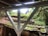 An old, weathered wooden roof is covered in green moss and small plants. A wooden structure is supported by white beams, with greenery and trees surrounding the area, contributing to a natural, overgrown appearance.