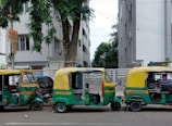 Three colorful auto rickshaws are parked in a row on the side of a street. Each has a green and yellow color scheme with decorative elements. The surroundings include tall residential buildings and lush green trees. A sign indicating a hostel is visible on the left side.
